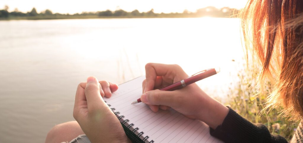 Woman writing in notebook next to lake during sunset